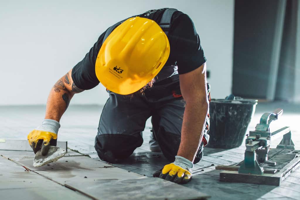 A bearded man in overalls working on a tile floor, filling the joints with grout.